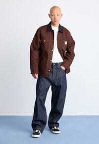 Brown canvas jacket with a dark collar, four pockets, and gold buttons, paired with loose, dark denim jeans and black sneakers with white accents.