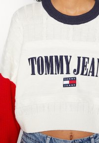 Pull court avec tricot en câble blanc et manches rouges. Il possède un col marine et le logo en texte "TOMMY JEANS" avec une étiquette de marque.