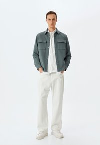 Grå button-up jakke med to brystlommer, båret over en hvid skjorte og parret med hvide bukser og sneakers. Glat stof og afslappet pasform.