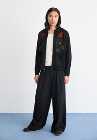 Juoda megzta kardiganas su oranžinės ir žalios gėlės siuvinėjimu, su užtrauktuku priekyje ir dviem priekinėmis kišenėmis. Sujungiama su laisvomis juodomis kelnių.