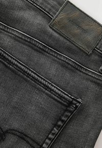 Gros plan d'un jean en denim gris foncé montrant la poche arrière et l'étiquette de marque en cuir cousue indiquant "Denim Supply."