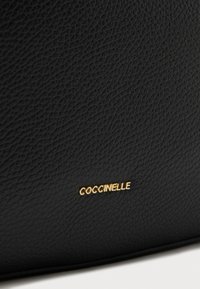 Borsa a mano in pelle nera testurizzata con un piccolo logo "Coccinelle" in oro. Il materiale ha una finitura pebbled, che ne esalta la qualità tattile.