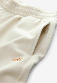 Svijetlosmeđi trenirka hlače izrađene od mekanog materijala, s elastičnim pojasom, bočnim džepovima i narančastim izvezenim "self" logom sprijeda.