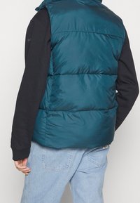 Türkiser Stepp-Weste mit Kapuze, horizontalen Nähten und einer glatten, glänzenden Oberfläche. Getragen über einem schwarzen Langarmshirt und hellblauen Jeans.