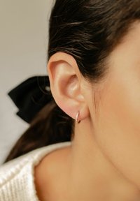 Boucles d'oreilles créoles en or rose avec pierres précieuses claires, présentées sur l'oreille d'un modèle. Cheveux attachés avec un ruban noir. Texture de peau douce visible.