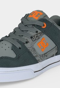 Baskets grises et noires avec logo DC orange sur la languette et le côté, lacets gris et semelle en caoutchouc blanche avec une fine bande brune.