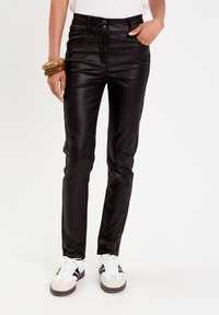 Pantalones ajustados de imitación cuero negro con cierre de dos botones y bolsillos frontales, combinados con zapatillas blancas y grises. Textura suave y brillante.