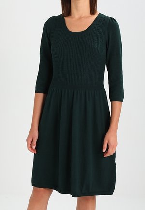 Vrouw draagt een donkergroene jurk tot op de knie met driekwart mouwen en een getextureerd gebreid lijfje.