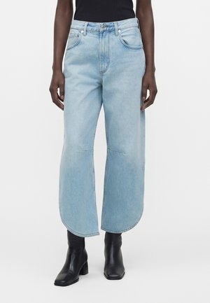Persona vistiendo vaqueros de pierna ancha azul claro con dobladillos redondeados, combinados con botines negros de tacón y una camiseta sin mangas negra.