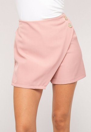 Shorts portefeuille rose clair avec deux boutons décoratifs beige sur le côté, portés avec un haut blanc.