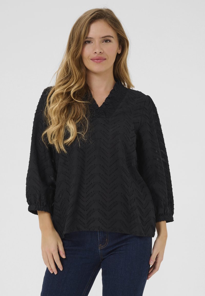 Blusa nera con scollo a V, maniche 3/4, motivo a zigzag testurizzato e dettagli arricciati al collo e ai polsi. Abbinata a jeans blu.