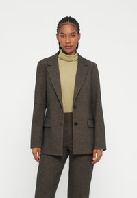 Rudas tweed blazeris su išpjautomis apykaklėmis, dviem priekinėmis kišenėmis ir vienu mygtuku, dėvimas ant šviesiai žalios turtlenecko.