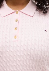 Κοντινή λήψη ενός κοντομάνικου πόλο πουλόβερ σε ανοιχτό ροζ χρώμα με πλεχτό σχέδιο καλωδίου, χρυσά κουμπιά και μικρό κεντημένο λογότυπο σε κόκκινο, λευκό και μπλε στο στήθος.