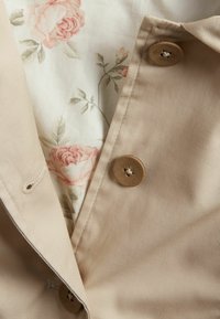 Beige stof met een gladde textuur, met zichtbare bruine knopen. Een bloemenpatroon voering in zachtroze en groen is gedeeltelijk zichtbaar.