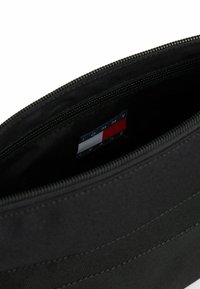 Pochette nera con zip e superficie testurizzata, con etichetta Tommy Jeans che presenta colori rosso, bianco e blu all'interno. Design minimalista.