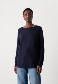 Mørkeblå langermet topp med vid hals, laget av et strukturert stoff, paret med lyseblå jeans. Enkelt og rent design.
