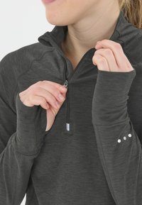 Person in einem dunkelgrauen, langärmligen Pullover mit Daumenlöchern, der den Reißverschluss vorne in der Nähe des Kragens hochzieht.