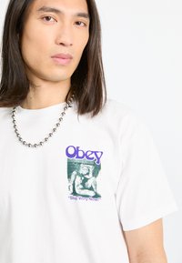 Hvit t-skjorte i bomull med grønn og svart grafikktrykk og lilla tekst "Obey" samt setningen "Stop Worry Now!!" på forsiden.