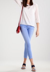 Jeans ajustados azul claro, blusa blanca con escote decorativo, combinados con sandalias negras y un bolso cruzado rojo.