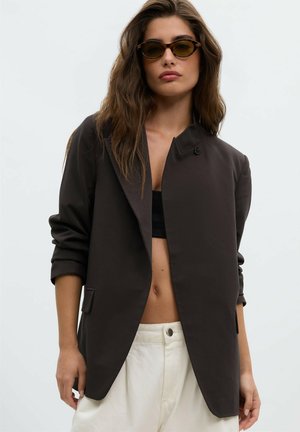 Femme portant un top court noir, un blazer sombre oversize, un pantalon écru et de petites lunettes de soleil foncées avec des cheveux ondulés sur un fond uni.