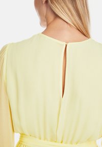 Gelbes Kleid aus strukturiertem Stoff, mit einem Rückenschlitz und gerafften Ärmeln. Weiches, fließendes Material.
