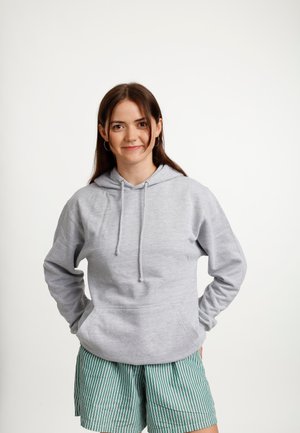 Młoda kobieta o brązowych włosach stoi uśmiechnięta, ubrana w jasnoszary kaptur i zielone spodenki w białe paski na jednolitym białym tle.