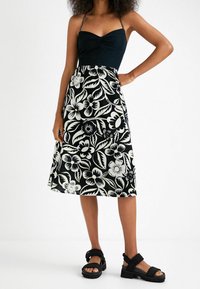 Vestido de patrón floral en blanco y negro con un cuerpo ajustado y falda acampanada, terminado con finas tirantes en los hombros; combinado con sandalias negras.