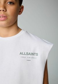 Biela bezrukávová tričko vyrobené z bavlny, s zeleným potlačeným textom "ALLSAINTS," s japonskými znakmi pod ním a "LONDON" nižšie.