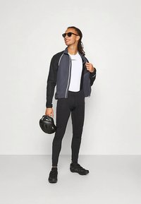 Sportieve fietsoutfit met een grijs en zwart zip-up jack, witte shirt, zwarte tights en zwarte schoenen. Houdt een zwarte helm vast.