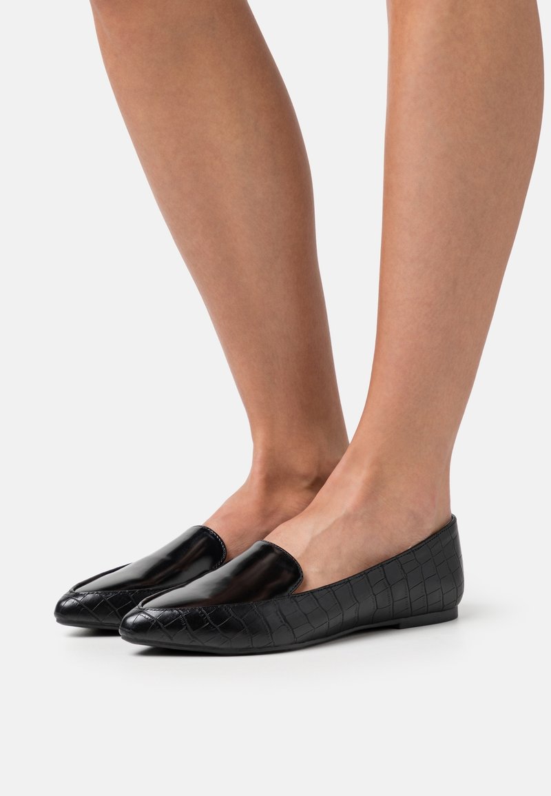 Jambes portant des mocassins plats noirs à motif crocodile avec des parties avant brillantes, debout sur un fond gris clair sans couture.