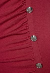 Rode textuur stof met verticale plooien aan de zijkant en vijf ronde zilveren knopen met reliëfdetail.