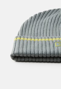 Szara, dziergana czapka typu beanie o prążkowanej teksturze, z żółtym paskiem i małą metką z logo naszytą z boku. Wykonana z miękkiego materiału przypominającego wełnę.