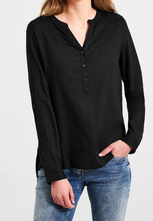 Frau trägt eine schwarze langärmelige Bluse mit Knöpfen und rundem Ausschnitt, kombiniert mit blauen Jeans, eine Hand in der Tasche vor schlichtem Hintergrund.