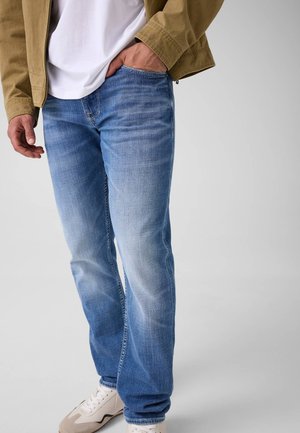 Mann trägt hellblaue ausgewaschene Jeans, weiße Sneakers, ein weißes Hemd und eine braune Jacke mit einer Hand in der Tasche vor einfarbigem Hintergrund.