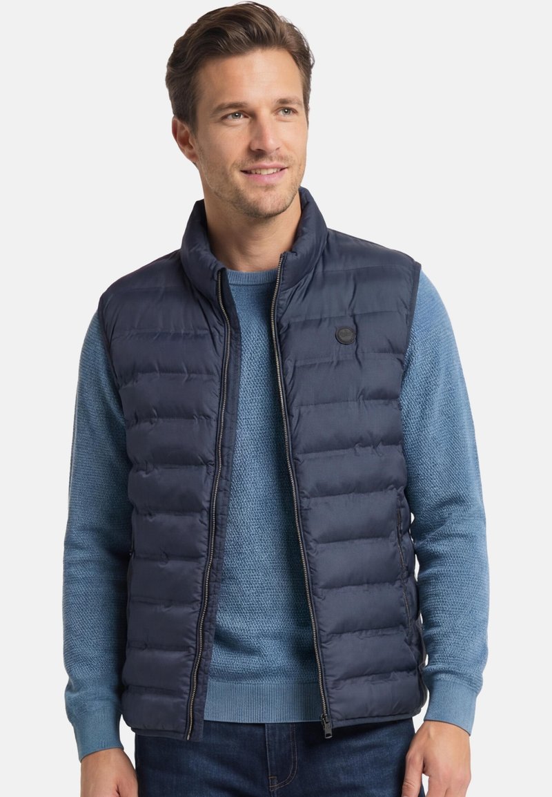 Man draagt een marineblauw doorgestikt vest over een lichtblauwe gebreide trui, staand en licht naar rechts kijkend met een subtiele glimlach.