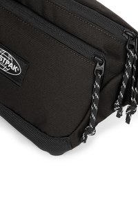 Pochette in tessuto nero con due scomparti chiusi da zip, esterno testurizzato e stringhe di chiusura in bianco e nero. Etichetta del logo visibile.