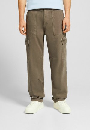Cargo-Hosen aus hellbraunem Stoff mit lockerer Passform, die mehrere Taschen aufweisen und mit weißen Sneakern kombiniert sind. Strukturiertes Material und sichtbare Nähte.