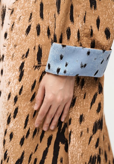 Χέρι που ακουμπά σε καφέ ύφασμα με μαύρα αφηρημένα στίγματα και μια γαλάζια μανσέτα διπλωμένη προς τα πάνω με αντίστοιχα μαύρα στίγματα.