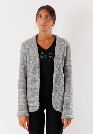 Blazer grigio in maglia con finitura testurizzata, con revers e apertura frontale. Indossato sopra un top nero a V e abbinato a pantaloni neri.