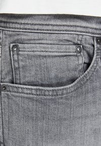 Jeans de mezclilla gris con un pequeño bolsillo frontal, que presentan herrajes de metal negro y costuras visibles a lo largo de los bordes. Detalle de tela texturizada visible.