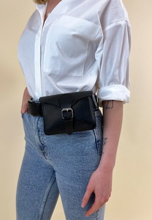 Personne portant un jean bleu, une chemise blanche à boutons avec les manches retroussées, et une ceinture-sacoche en cuir noir avec une boucle autour de la taille.