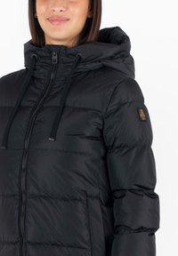 Giacca nera imbottita con cappuccio, dotata di una cerniera frontale, lacci regolabili e una patch decorativa sul braccio sinistro. Texture liscia e lucida.