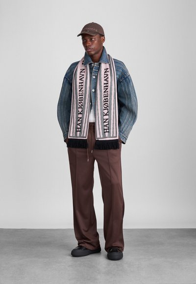 Džinsu jaka, balta t-kreklā, brūni bikses, melnas kurpes, rozā frēzētā šalle ar tekstu "HANK KØBENHAVN", brūns cepures ar logo.