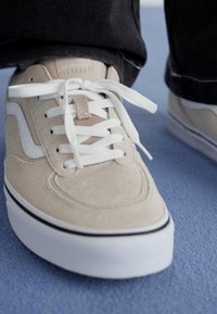 Beige suede sneaker with white accents and laces, featuring a cushioned sole and black trim. Visible stitching details are present.