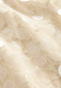 Lichtbeige stof met een doorschijnende textuur, versierd met verspreide grote, ronde pailletten die met fijne draadjes zijn verbonden, wat een delicaat patroon creëert.