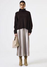 Brauner Rollkragenpullover, langes Kleid aus satinisiertem Silber, beiger Handtasche mit Ausschnittgriff und beigefarbenen High Heels. Einheitlicher weißer Hintergrund.