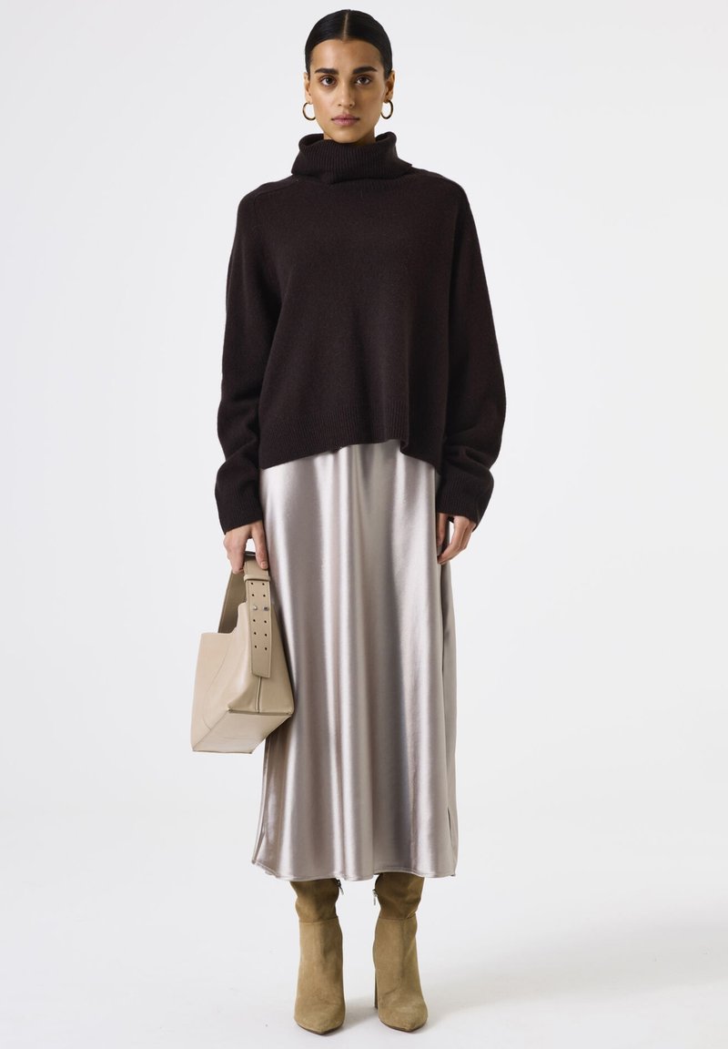 Brauner Rollkragenpullover, langes Kleid aus satinisiertem Silber, beiger Handtasche mit Ausschnittgriff und beigefarbenen High Heels. Einheitlicher weißer Hintergrund.