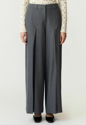 Pantalon gris à jambes larges avec plis à l'avant, porté avec un haut en dentelle crème à manches longues et des chaussures noires pointues.