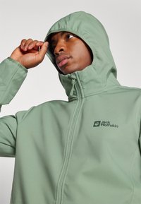 Green zip-up jacket featuring a hood, soft texture, and the Jack Wolfskin logo on the chest. Seam detailing adds structure to the design.