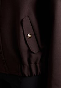 Brun jakke med struktureret stof, en klaplomme med en gylden knap og detaljer med samlet kant. Nærbillede af ærmeområdet.